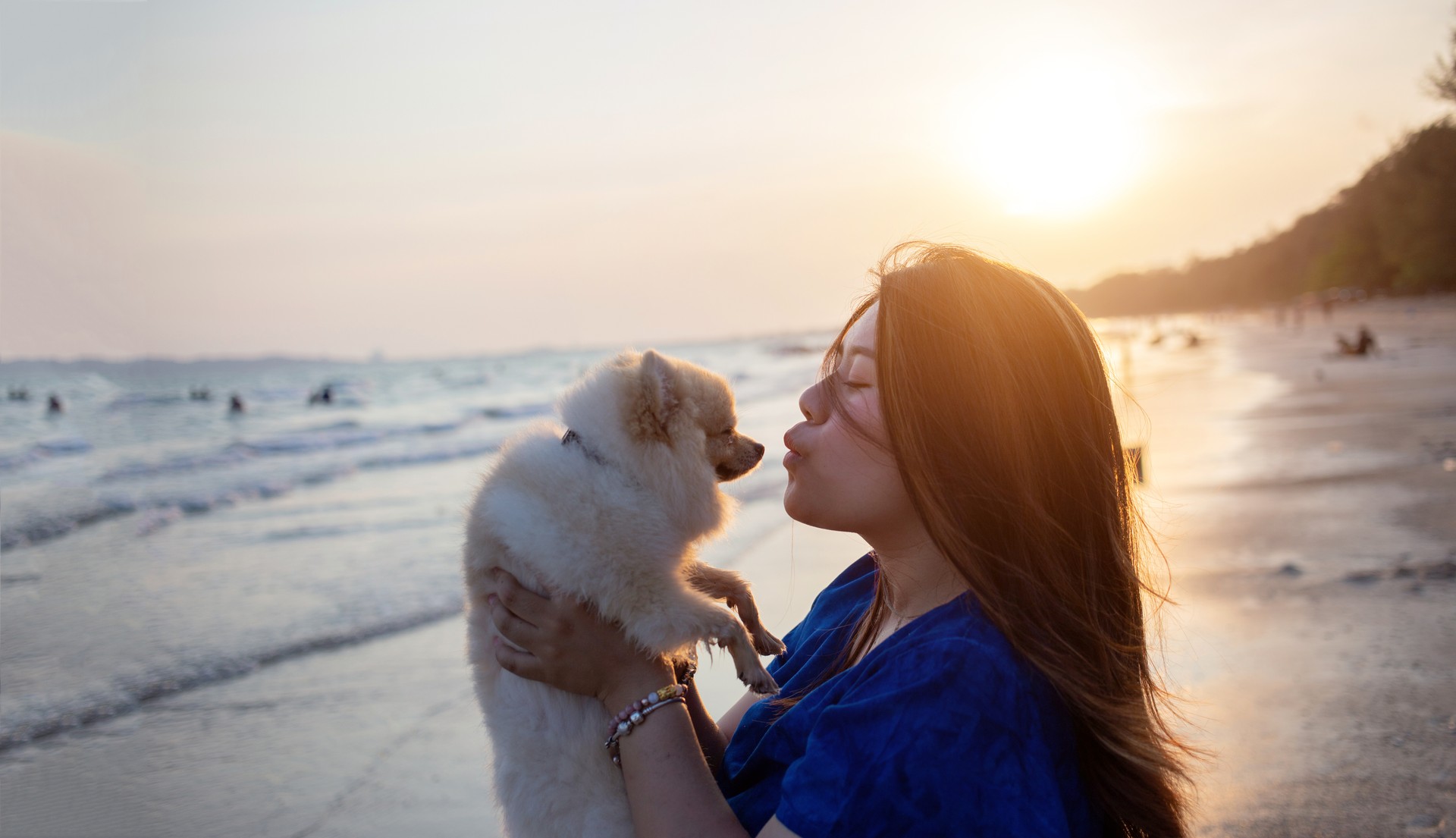 Young Woman Holding Dog While Standing At Beach During Sunset